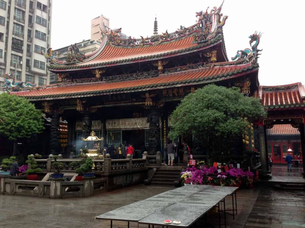 Longshan temple in Taipei, Taiwan. Asian temple with double level roof. Trees on left and right of the temple entrance. Grey tables in front of the trees.